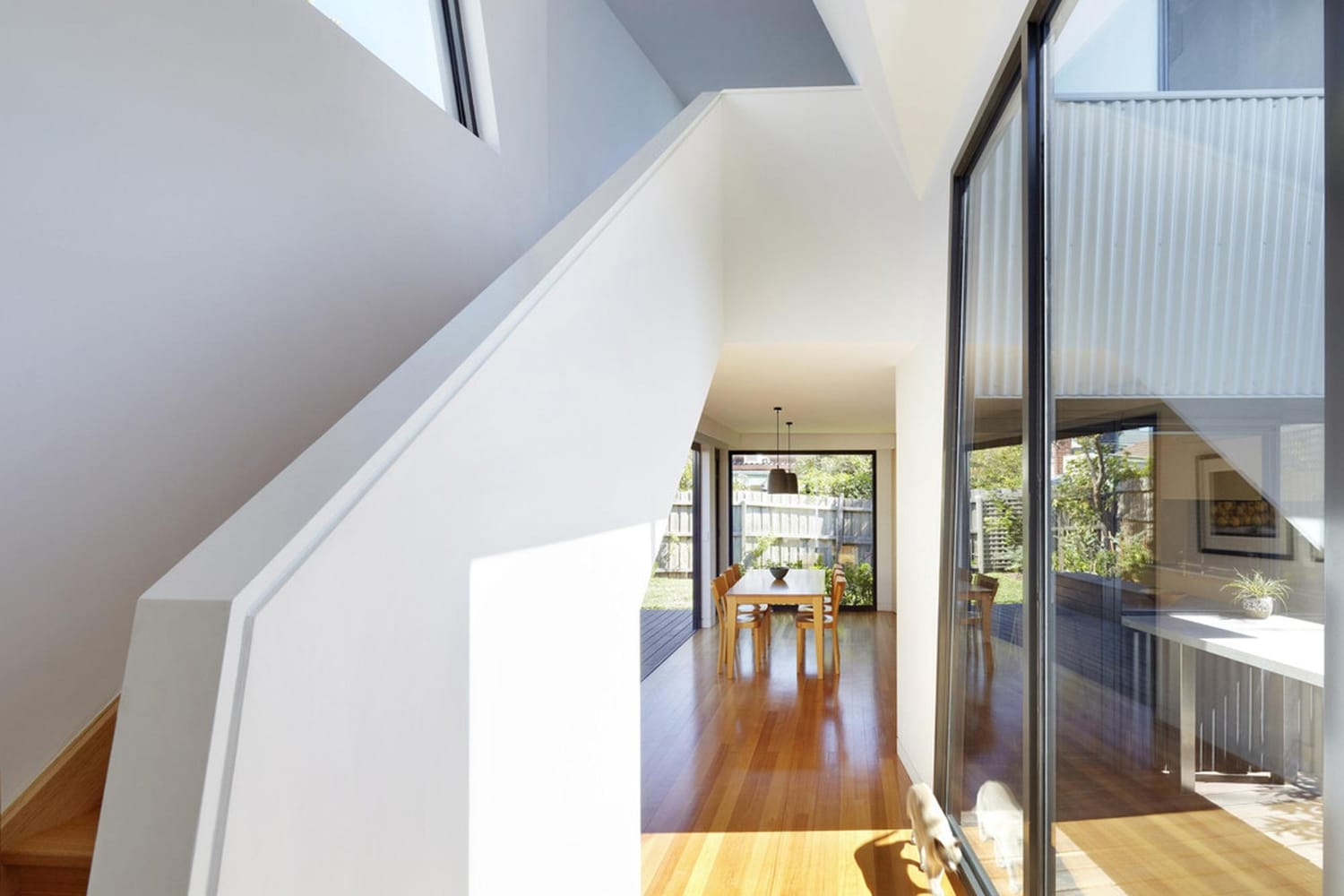 Bright modern hallway with wooden floor, white walls, and large windows, leading to a dining area with a table.